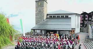 Lebendige Traditionen. Geistliche, Musikanten und Ehrenkompanie versammeln sich in Reih und Glied vor der Kirche von Visperterminen.